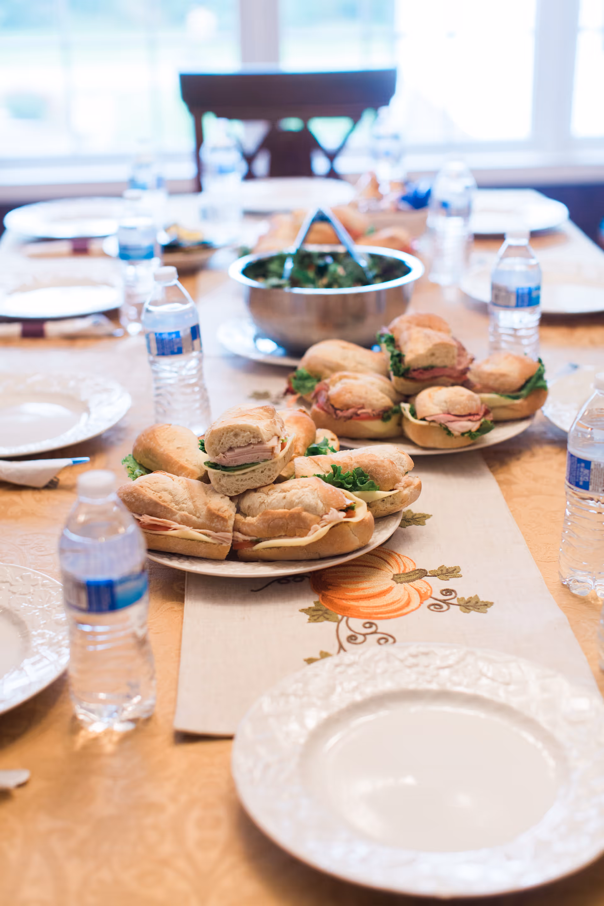 A dining table set with plates, bottled water, and plates of sandwiches with lettuce and deli meat. In the background, there is a bowl of salad with serving tongs. The table has a decorative table runner with a pumpkin design.