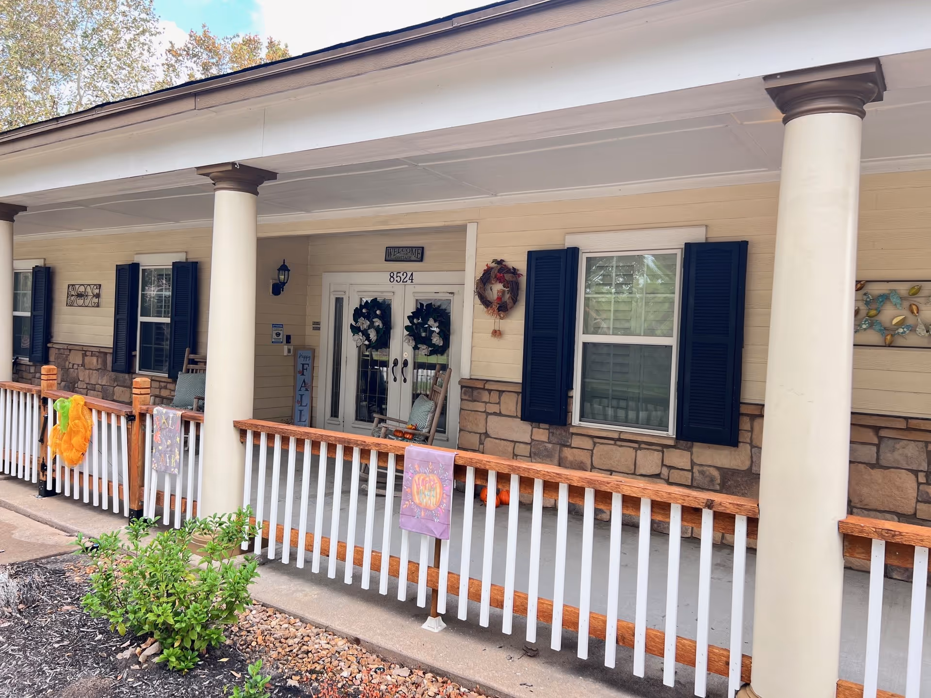 Front porch of a building with beige siding and stone accents, featuring white columns and a wooden railing decorated with fall-themed banners. Two wreaths hang on the double glass doors, which have the number 8524 above them. There are windows with dark blue shutters on either side of the doors, and a small garden with green plants in front of the porch.