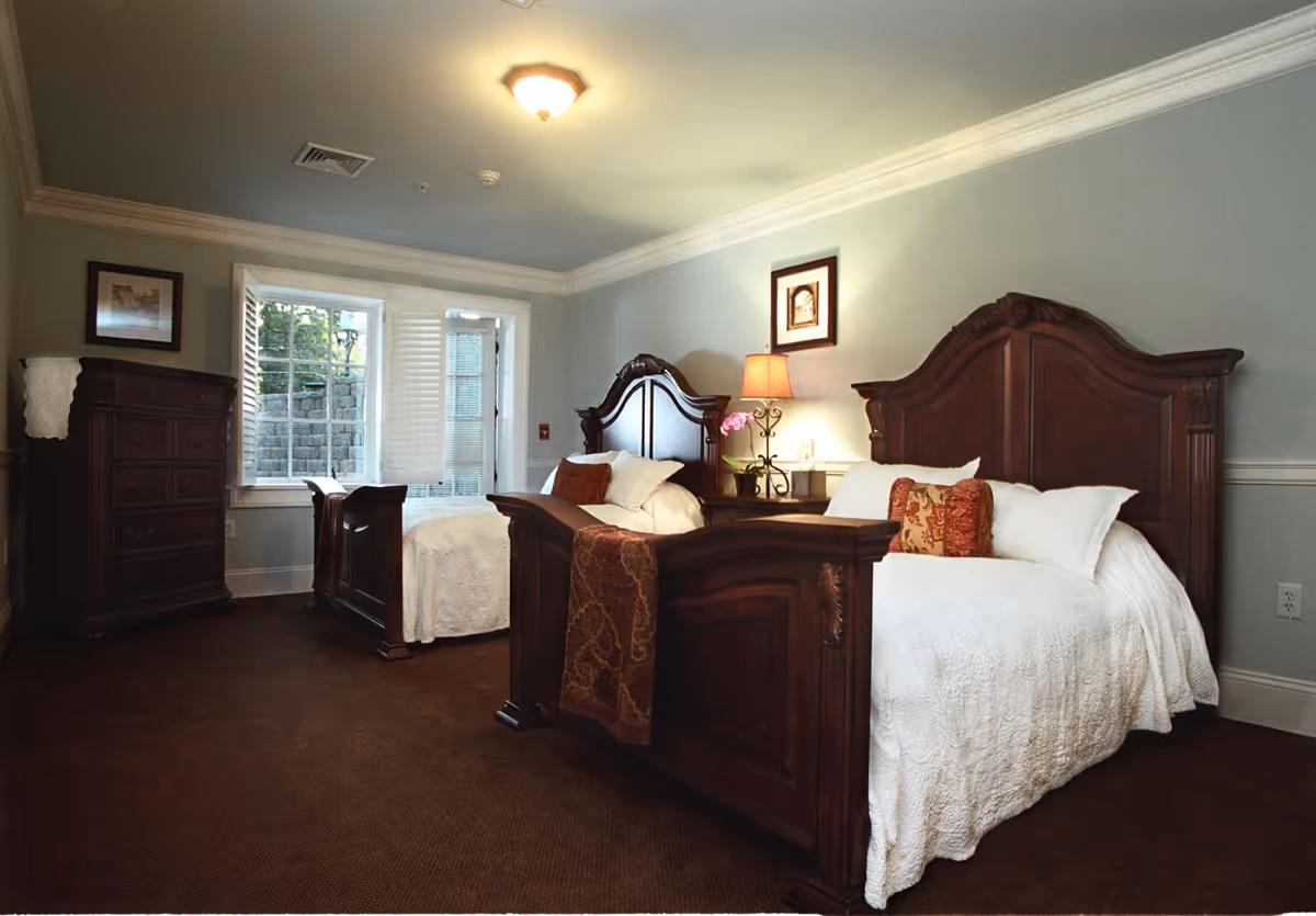 A bedroom with two wooden twin beds featuring ornate headboards and footboards. Each bed is made with white bedding and decorative pillows. Between the beds is a nightstand with a lamp and a small potted plant. There is a wooden dresser on the left side of the room, a window with white shutters, and a door leading outside. The walls are painted light blue with white trim, and the floor is carpeted in brown.