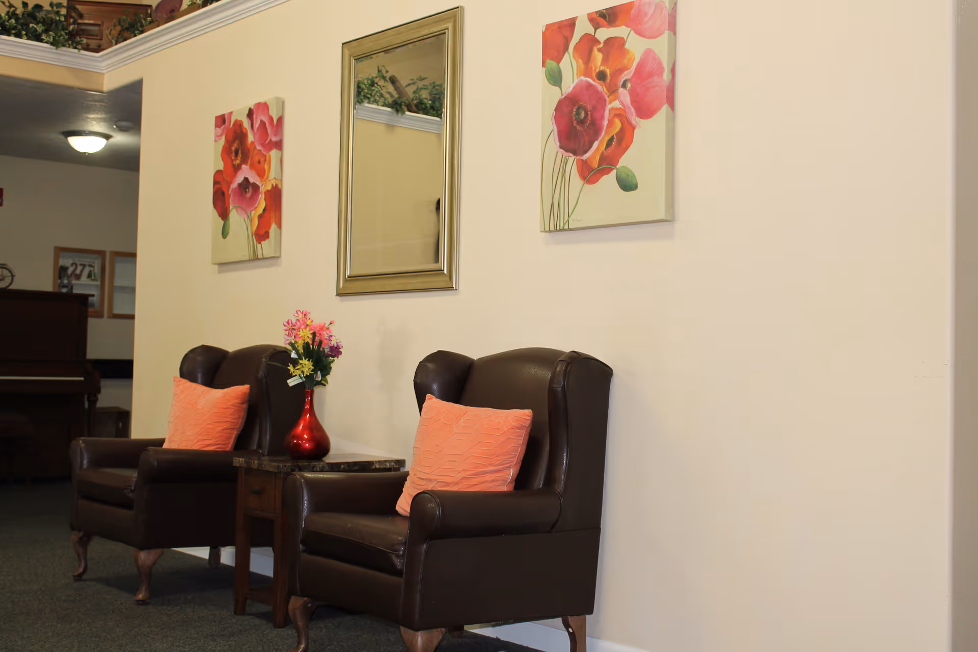 Two dark brown leather armchairs with coral-colored pillows are placed against a beige wall. Between the chairs is a small wooden side table with a red vase holding colorful flowers. Above the chairs, there is a rectangular mirror flanked by two floral paintings featuring red and pink flowers. In the background, a piano and some framed pictures are partially visible.