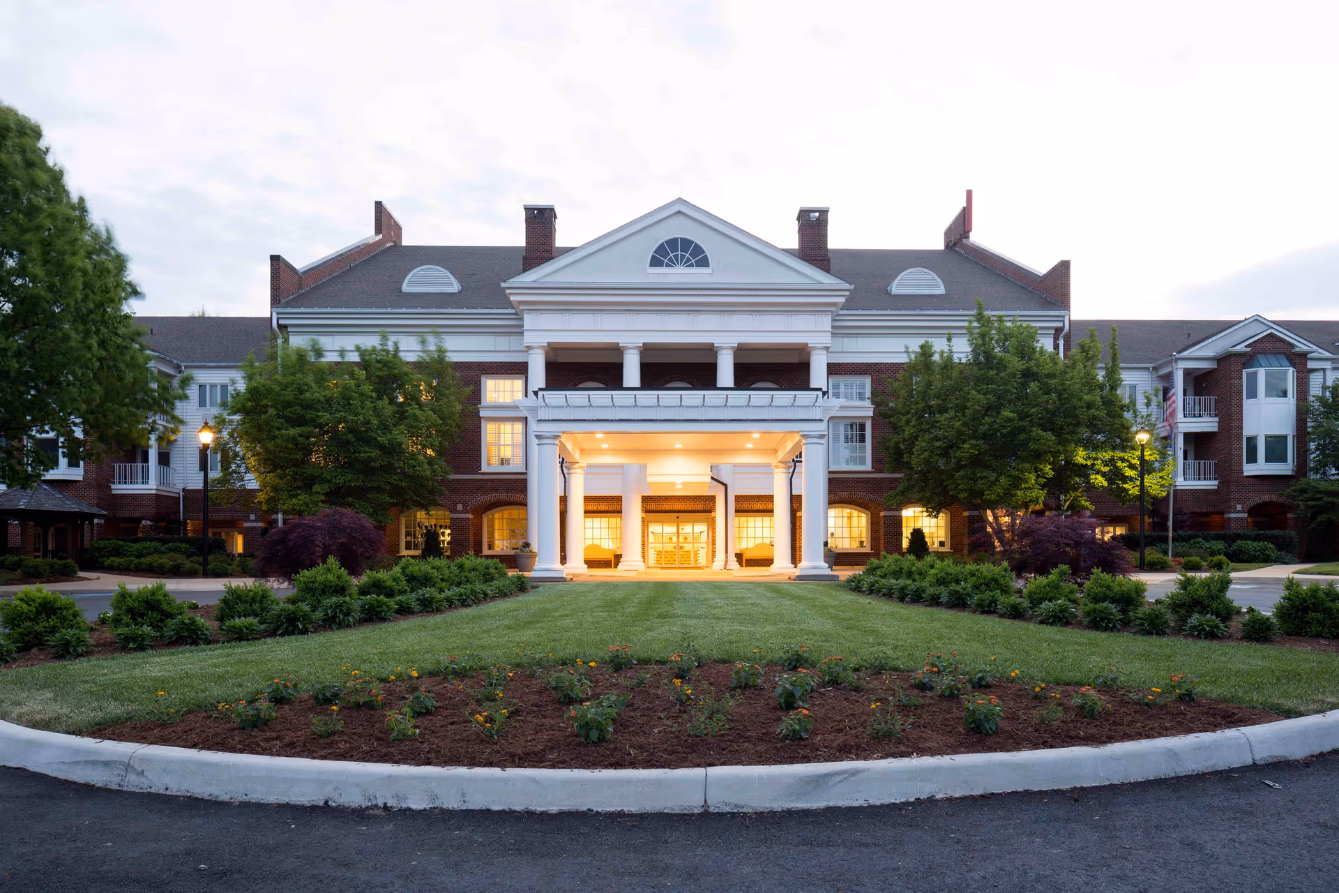 Front exterior view of a large, elegant senior living facility building with a well-maintained lawn and landscaping. The building features a prominent white columned entrance with warm lighting inside, red brick walls, and multiple windows. Trees and shrubs flank the entrance area.