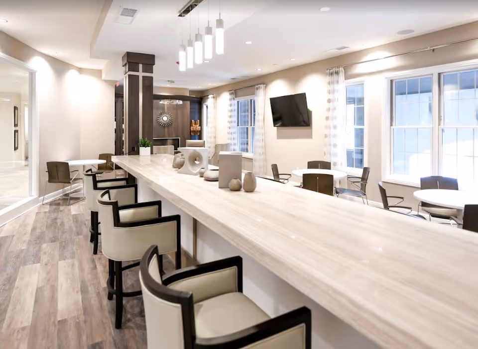 Bright communal dining and lounge area with a long bar counter, upholstered stools, round tables, and large windows.
