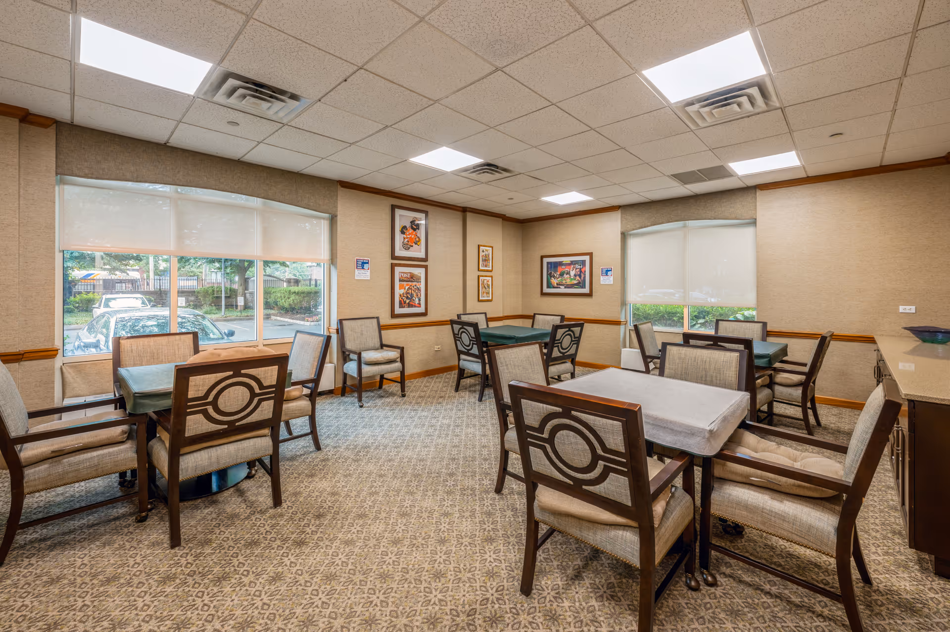 A well-lit room with multiple tables and chairs arranged for group seating, featuring large windows with roller shades partially down, framed artwork on the walls, and a patterned carpeted floor.