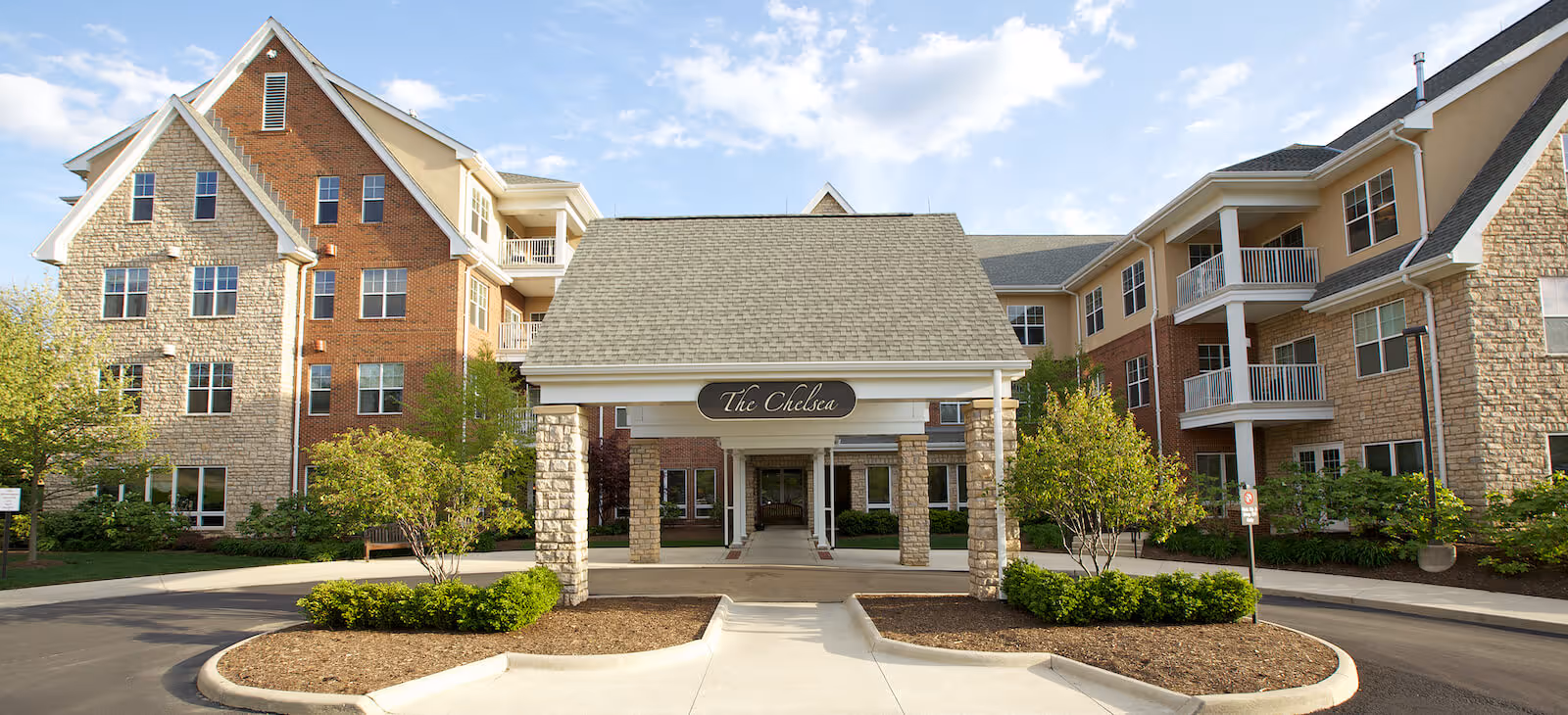 Front exterior view of a senior living facility named The Chelsea, featuring a covered entrance with stone pillars, surrounded by landscaped greenery and a multi-story building with brick and stone facade under a partly cloudy sky.