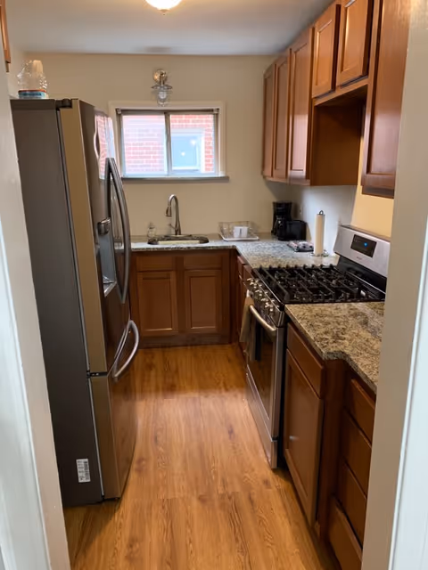 A narrow kitchen with wooden cabinets, granite countertops, a stainless steel refrigerator on the left, a gas stove with oven on the right, a sink under a small window, and a coffee maker on the countertop.