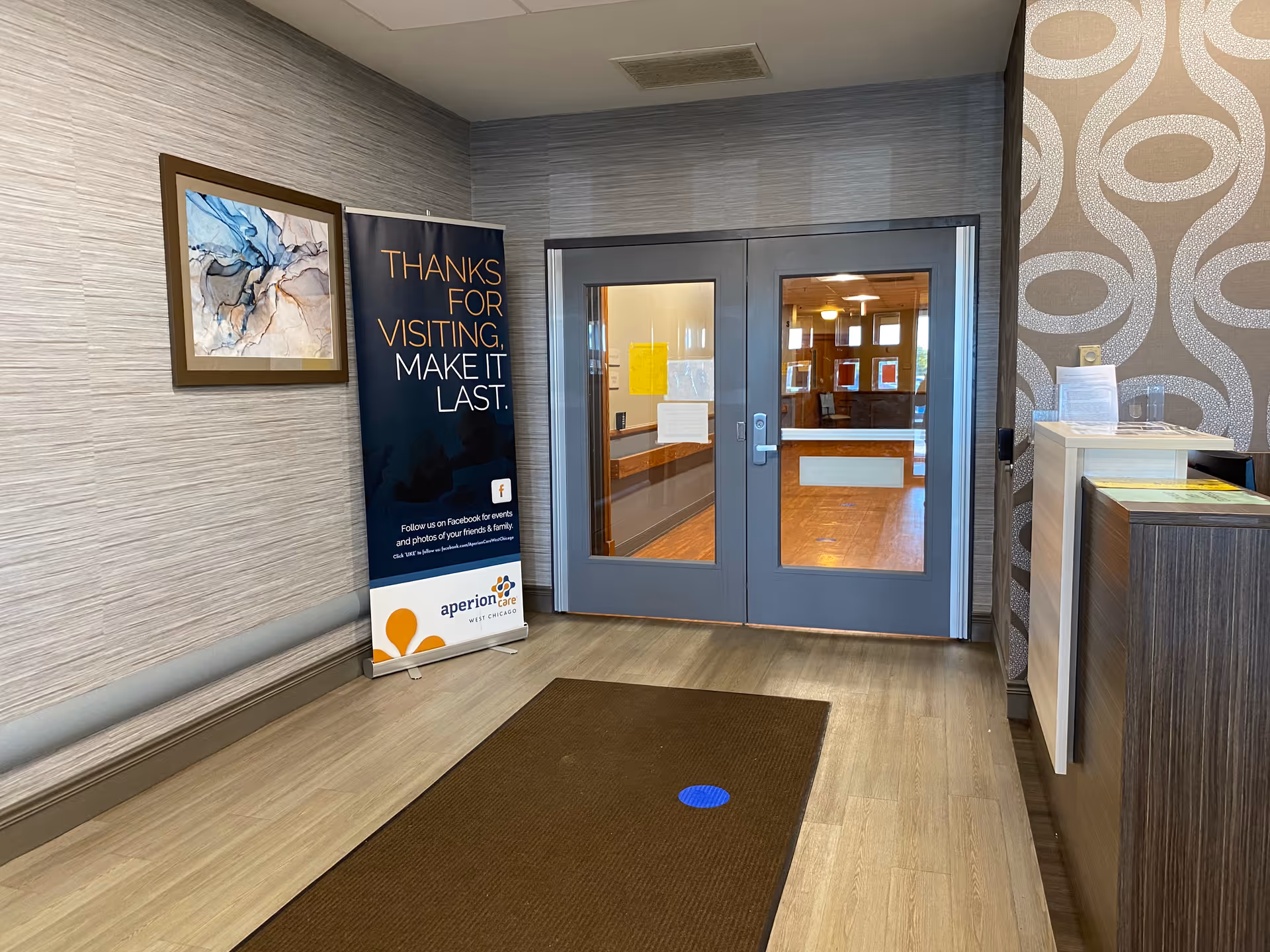 Interior view of a facility entrance area with double glass doors leading to a room with wooden flooring. To the left of the doors is a standing banner that reads 'Thanks for visiting, make it last.' There is a framed abstract artwork on the wall above the banner. On the right side, there is a reception desk with some papers on it. The floor has a brown mat with a blue circular sticker.