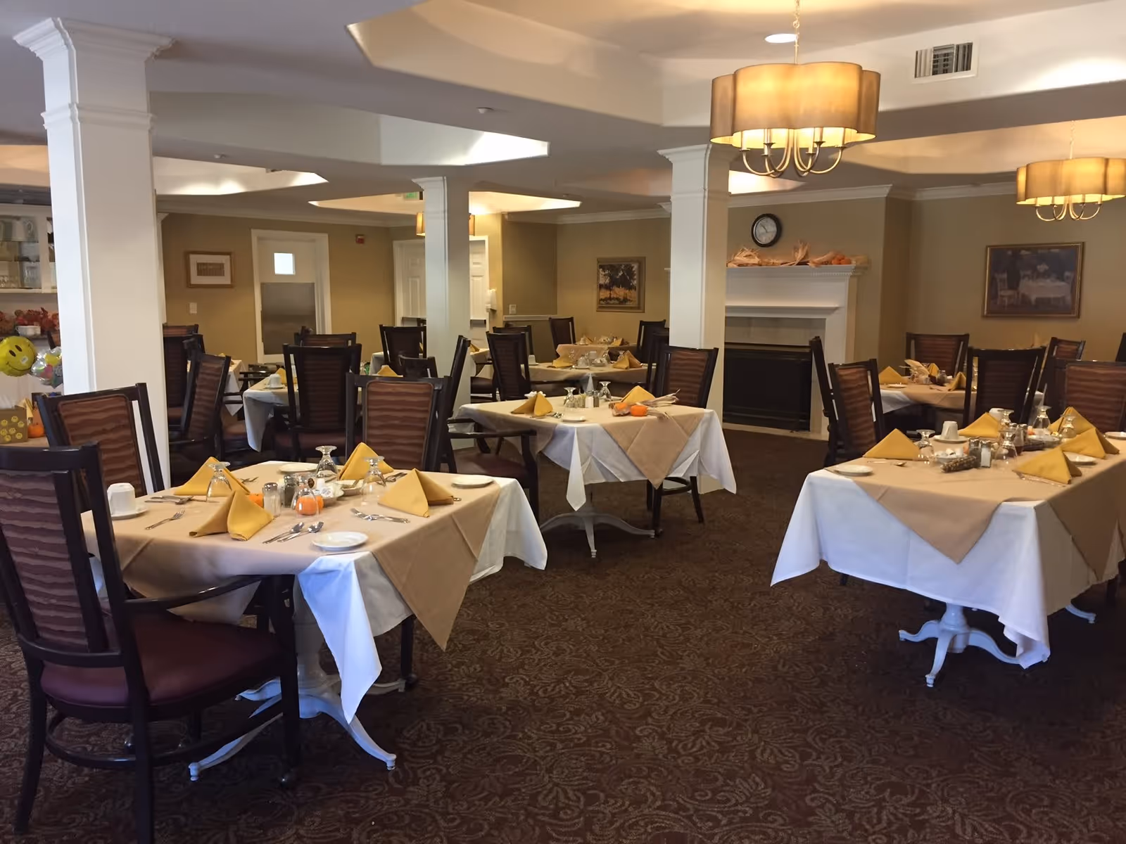 A dining room with multiple tables covered in white and beige tablecloths, each set with yellow folded napkins, plates, glasses, and silverware. The room has brown patterned carpet, beige walls, white pillars, and two chandeliers hanging from the ceiling. There is a fireplace with decorative items on the mantel and framed pictures on the walls.