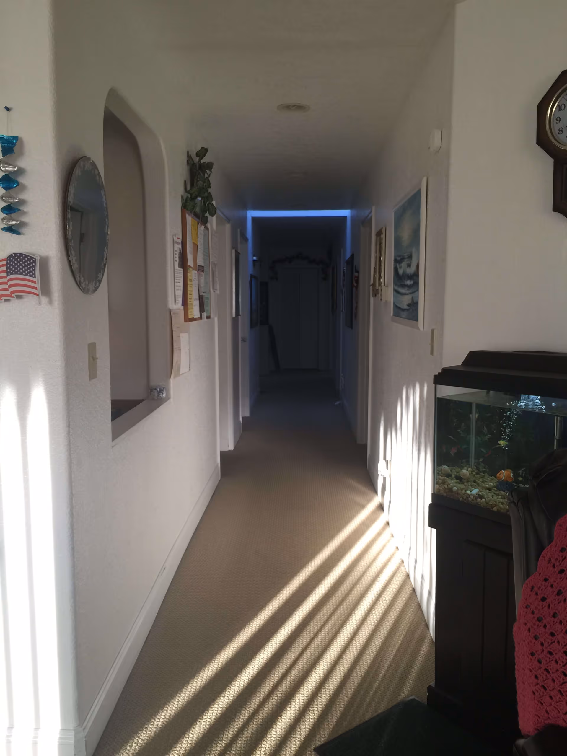 A long, narrow hallway in a residential or care home setting with white walls and beige carpet. On the left wall, there is a round mirror, an American flag decoration, and a bulletin board with papers pinned to it. On the right side, there is a framed painting of waves, a wall clock, and a fish tank with colorful fish. Sunlight casts striped shadows on the carpet from a window or blinds.