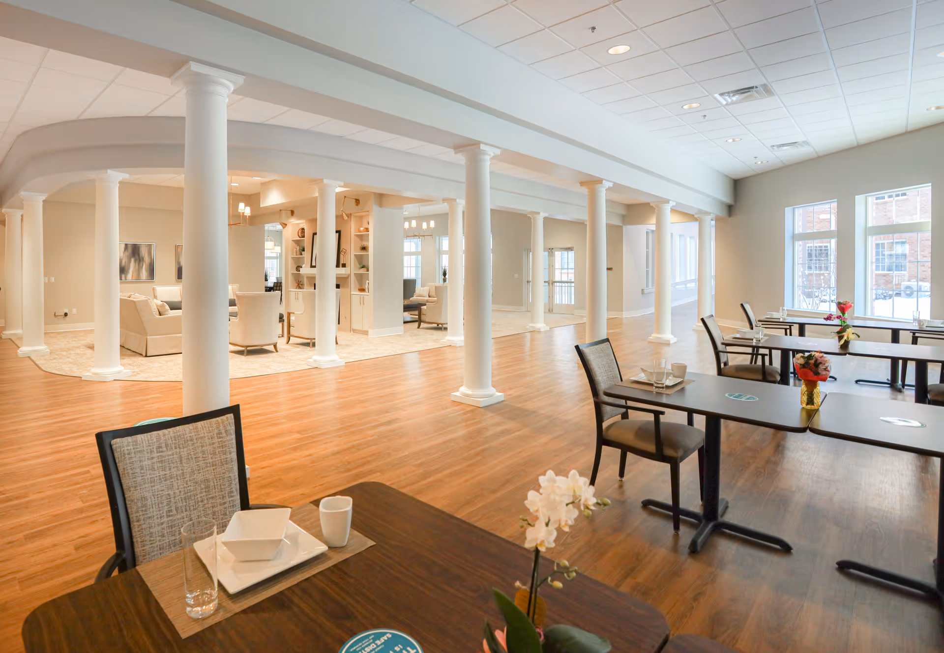 Spacious senior living dining area with wooden floors, multiple dining tables and chairs, white columns, and a lounge area in the background.
