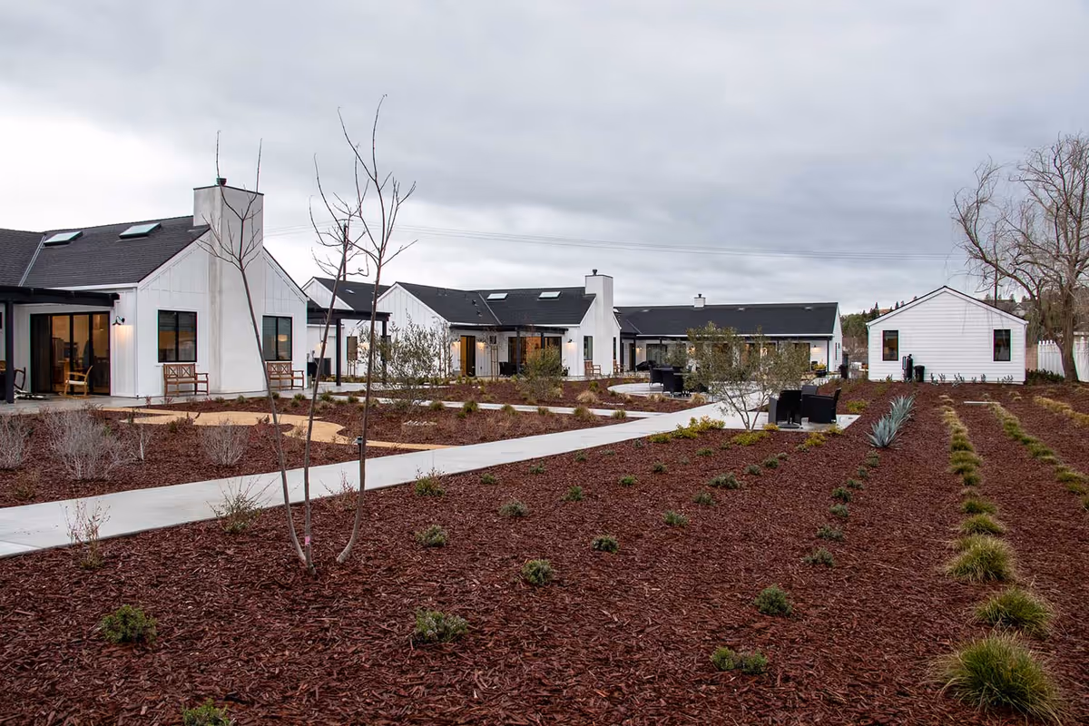 Outdoor view of a senior living facility named A Heavenly Home featuring white buildings with black roofs, a paved walkway, landscaped garden beds with mulch, small plants, and leafless trees under a cloudy sky.