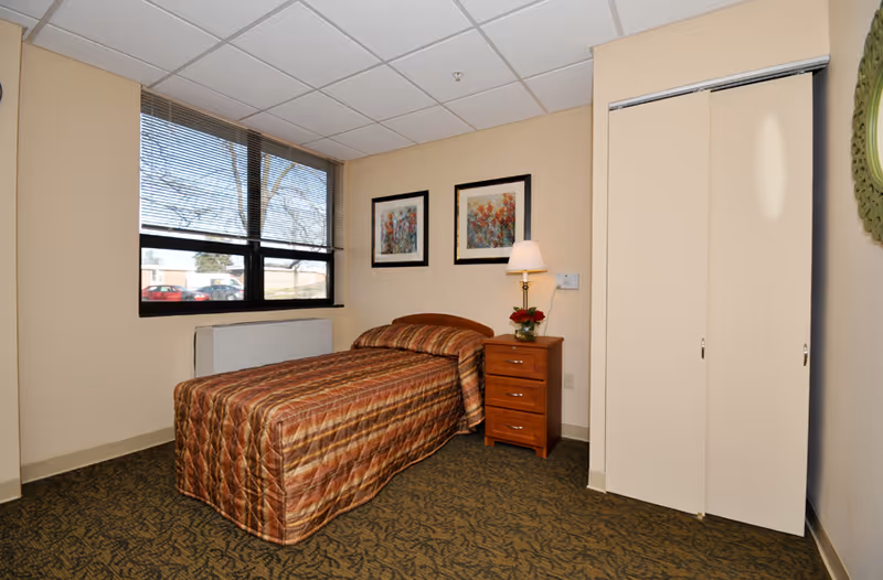 A small single bedroom with a twin bed, nightstand and lamp, framed artwork on the wall, a window, and a closet.