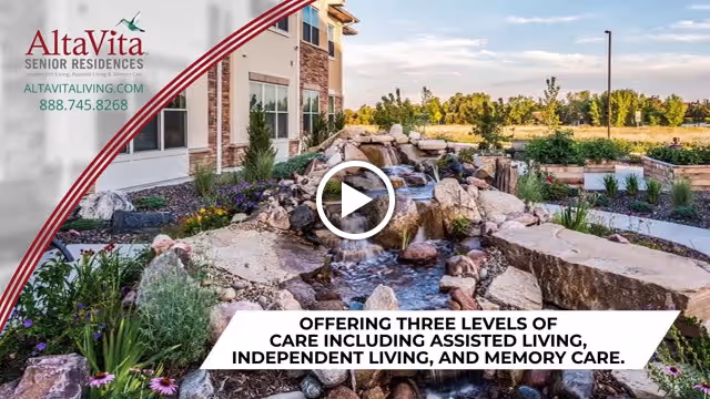 Outdoor garden area at AltaVita Memory Centre featuring a rock waterfall surrounded by plants and flowers, with a building and open landscape in the background under a partly cloudy sky.