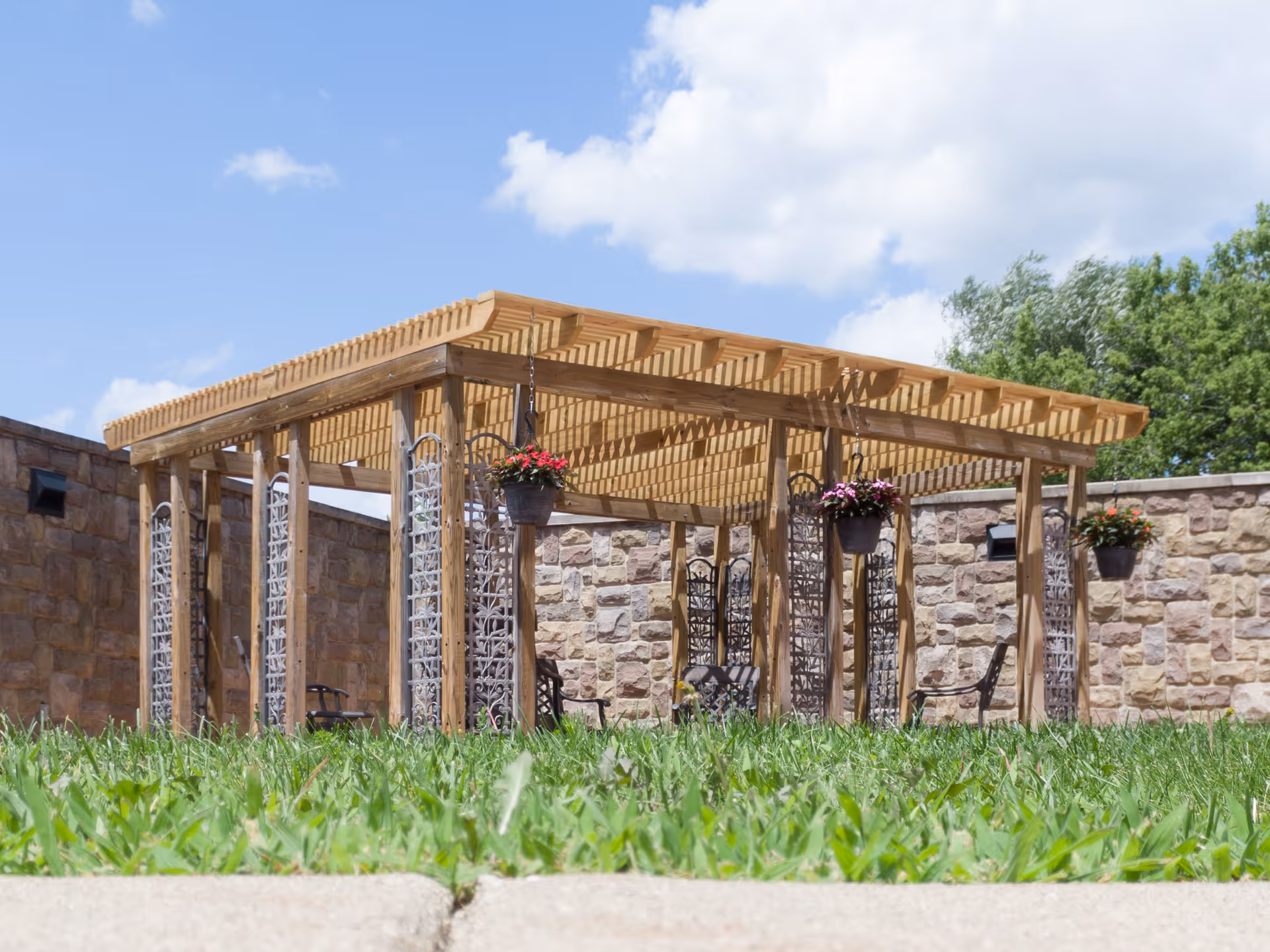Outdoor wooden pergola structure with hanging flower pots and metal chairs, surrounded by stone walls and green grass under a blue sky with some clouds.