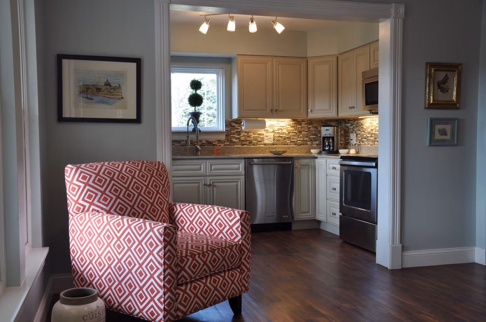 Interior view of a living space showing a red and white patterned armchair in the foreground and a kitchen area in the background with white cabinets, stainless steel appliances, a mosaic tile backsplash, and a window above the sink.