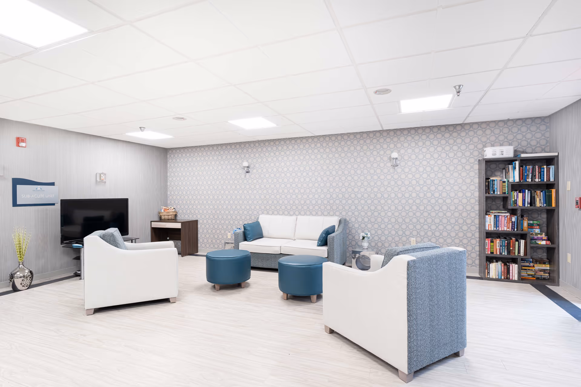 A bright and modern lounge area with light wood flooring, a patterned accent wall, and a white ceiling with recessed lighting. The room features a white and blue sofa, two matching armchairs, two blue round ottomans, a small side table with a flower vase, a bookshelf filled with books, a flat-screen TV mounted on a stand, and a small cabinet with a basket on top. A sign on the wall reads 'SUB-ACUTE UNIT'.