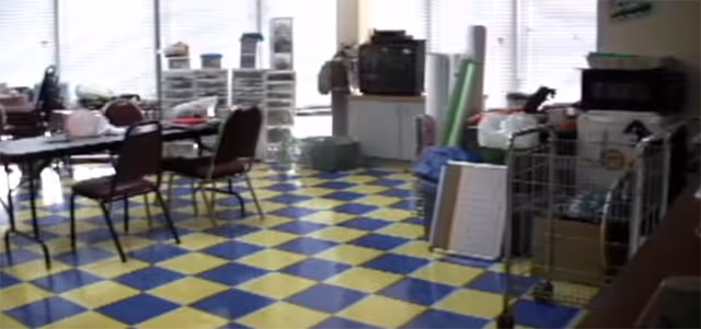 Interior activity/common room with blue-and-yellow checkered floor, folding tables and chairs, and storage shelves by windows.