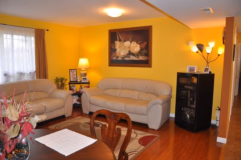 A cozy living room with yellow walls, featuring two beige leather sofas, a wooden coffee table with a white cloth, a wooden dining chair, a floor lamp with multiple lights, a framed floral painting on the wall, and a small black cabinet with family photos on top. There is a window with sheer white curtains and brown drapes, and a decorative plant on a side table.