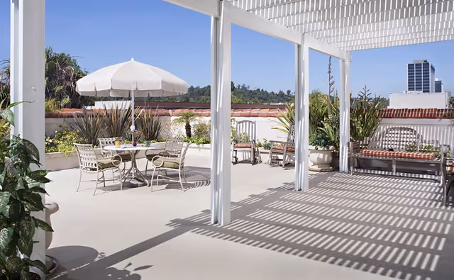Sunlit rooftop terrace with a pergola casting shadows, a table with umbrella and chairs, benches and potted plants overlooking distant buildings.