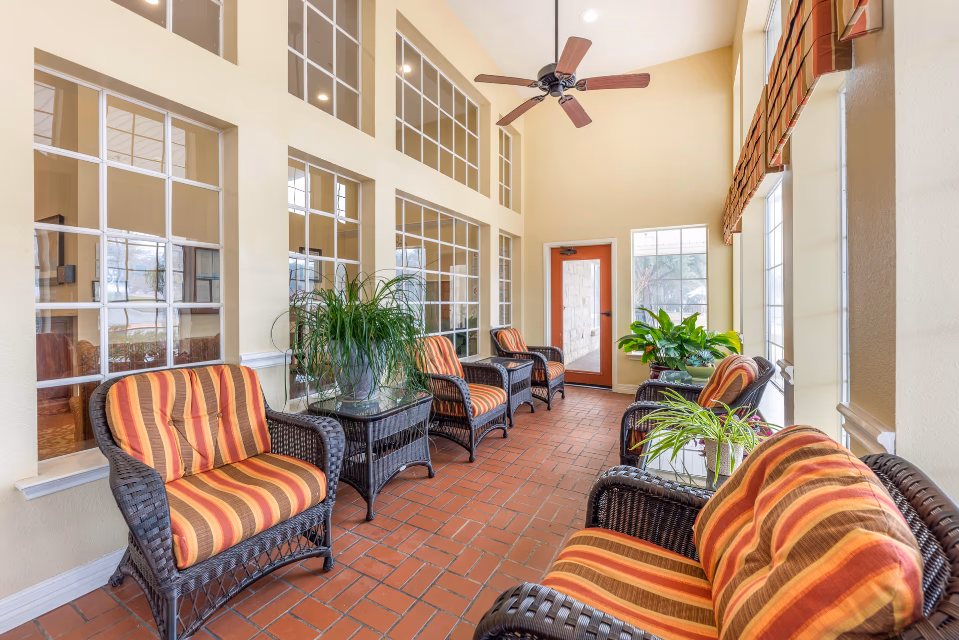 A bright indoor seating area with wicker chairs and loveseats featuring orange and red striped cushions. The floor is covered with reddish-brown tiles, and there are several green potted plants on glass-top tables between the chairs. Large windows and glass doors allow natural light to fill the space, and a ceiling fan is mounted on the high ceiling.