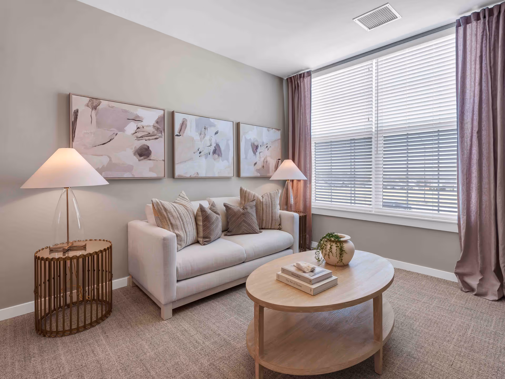 A cozy living room with a light beige sofa adorned with multiple textured pillows. Above the sofa, there are three abstract paintings in soft pastel tones. On either side of the sofa are modern table lamps with white shades, placed on round side tables with a gold metal frame. In front of the sofa is a light wooden oval coffee table with a small plant in a beige pot and a few stacked books. The room has a large window with white blinds and mauve curtains, and the floor is carpeted in a neutral tone.