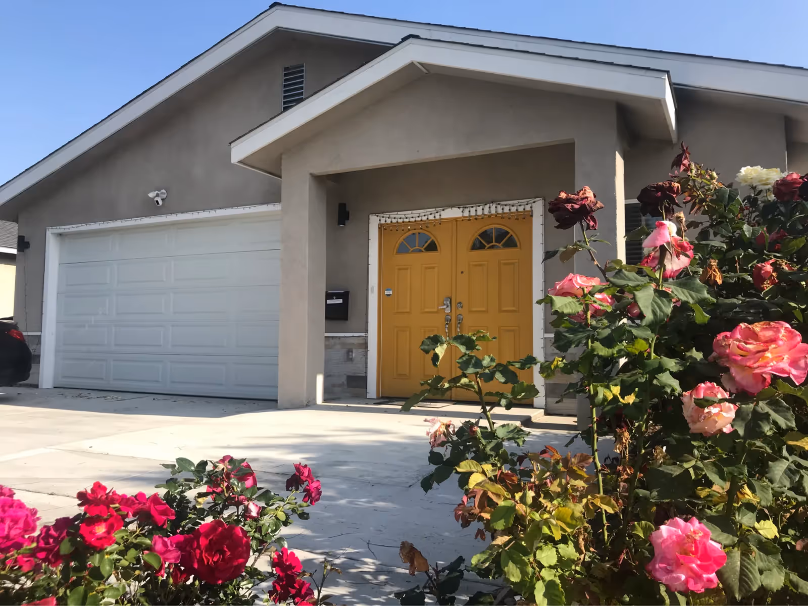 Front exterior view of a beige building with a double yellow door entrance and a white garage door. There are blooming pink and red roses in the foreground under a clear blue sky.