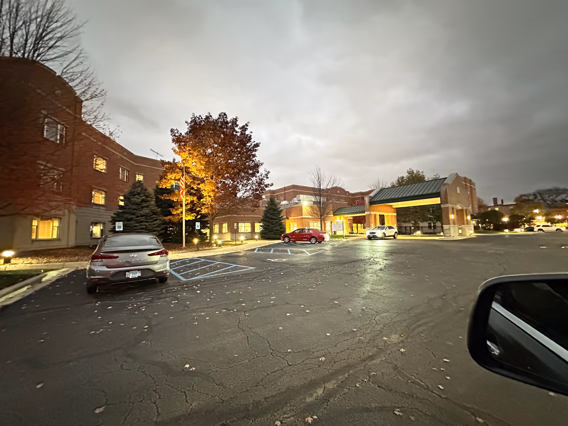 Exterior view of a brick senior living facility entrance and parking lot at dusk with parked cars and illuminated windows.