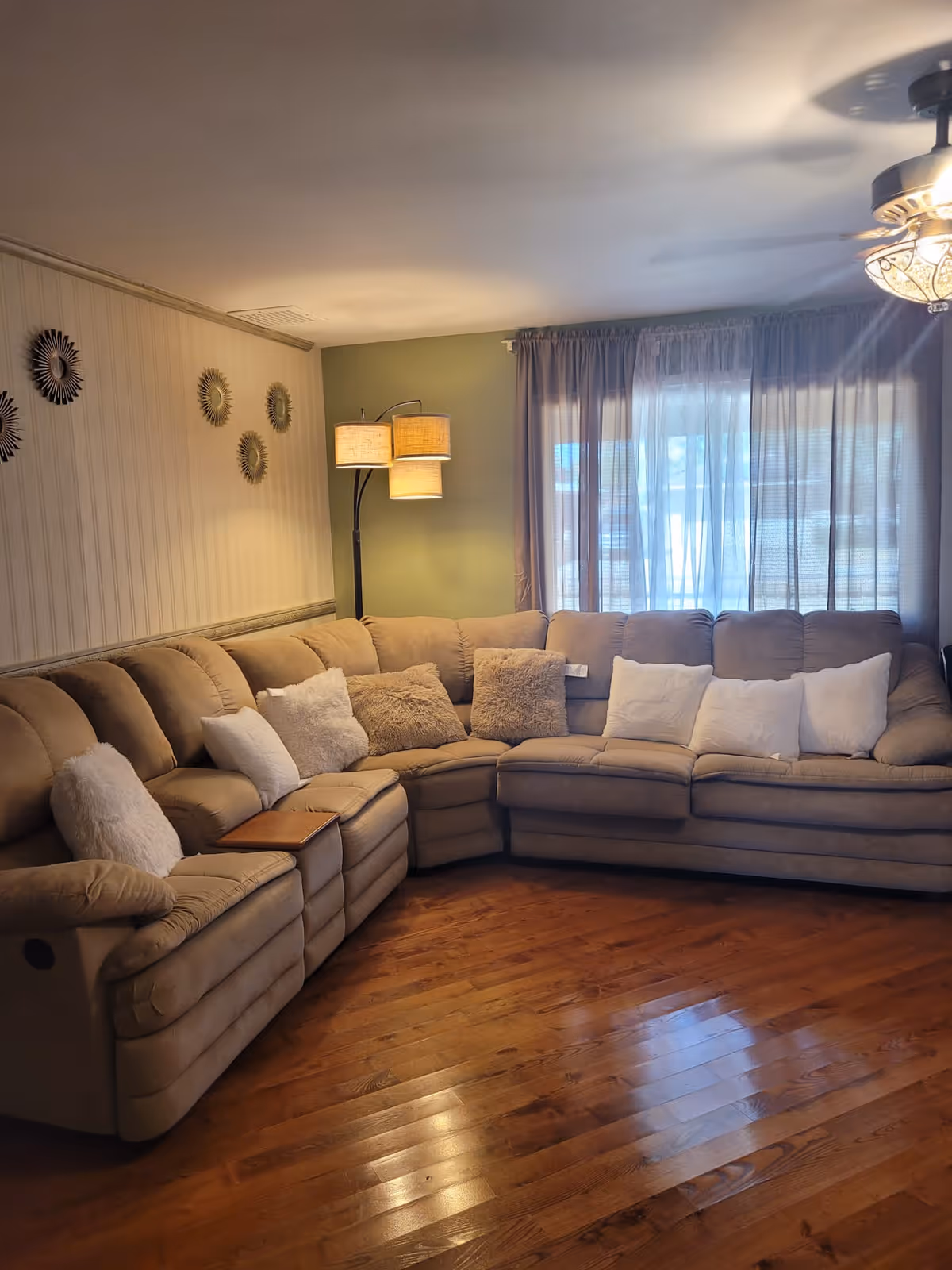 A cozy living room with a large beige sectional sofa adorned with multiple white and beige throw pillows. The room has wooden flooring, a green accent wall with a floor lamp featuring two lampshades, and a window covered with sheer curtains allowing natural light to filter in. Decorative sunburst wall hangings are displayed on a beige wall to the left.