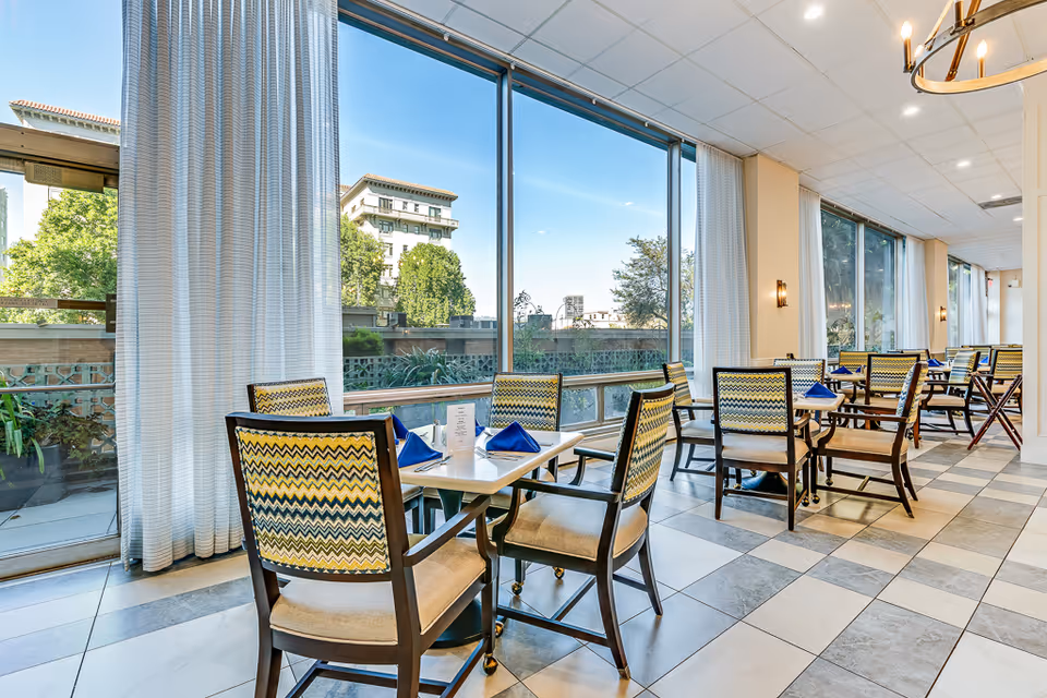A bright dining room with large windows showing an outdoor view of trees and buildings. The room has several tables with chairs featuring patterned backs and beige cushions. Each table is set with blue folded napkins and menus. The floor is tiled in a checkered pattern, and sheer white curtains hang beside the windows.