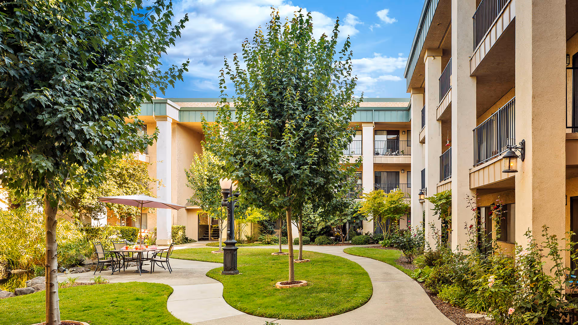 Outdoor courtyard area of a senior living facility with a curved concrete walkway, green grass, several trees, outdoor seating with a table and umbrella, and a multi-story building with balconies surrounding the courtyard.