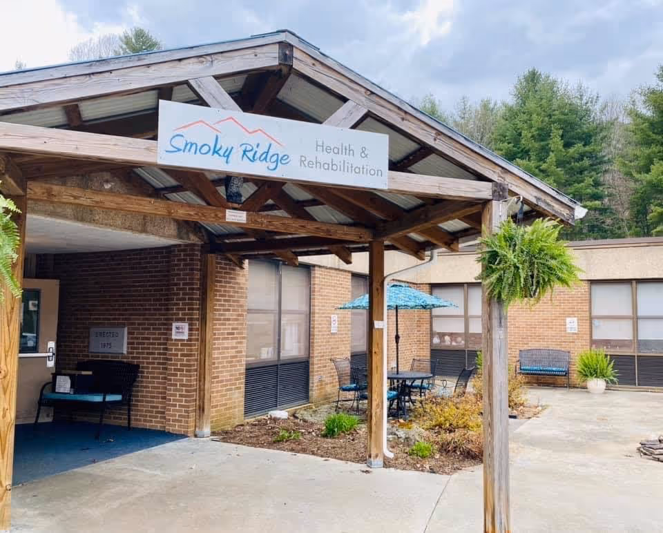 Covered wooden entrance canopy with a 'Smoky Ridge Health & Rehabilitation' sign above a courtyard with patio seating and plants in front of a brick building.