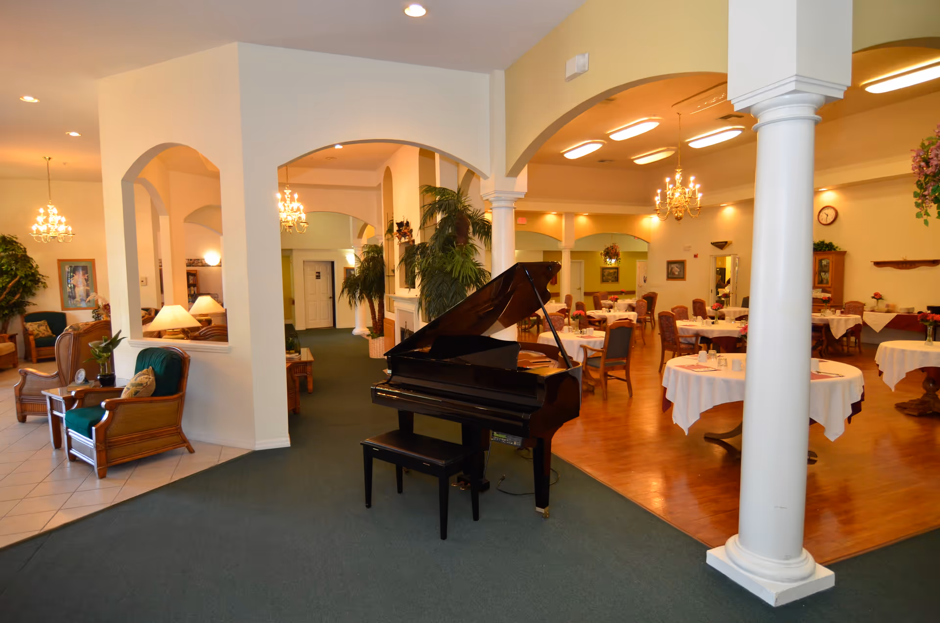 Interior view of a senior living facility featuring a black grand piano with a bench in the center. To the right, there is a dining area with round tables covered in white tablecloths and set with cups and napkins. The room has wooden flooring and is illuminated by chandeliers. To the left, there is a seating area with wicker chairs and green cushions, lamps, and plants. The space is decorated with arches, columns, and warm lighting.