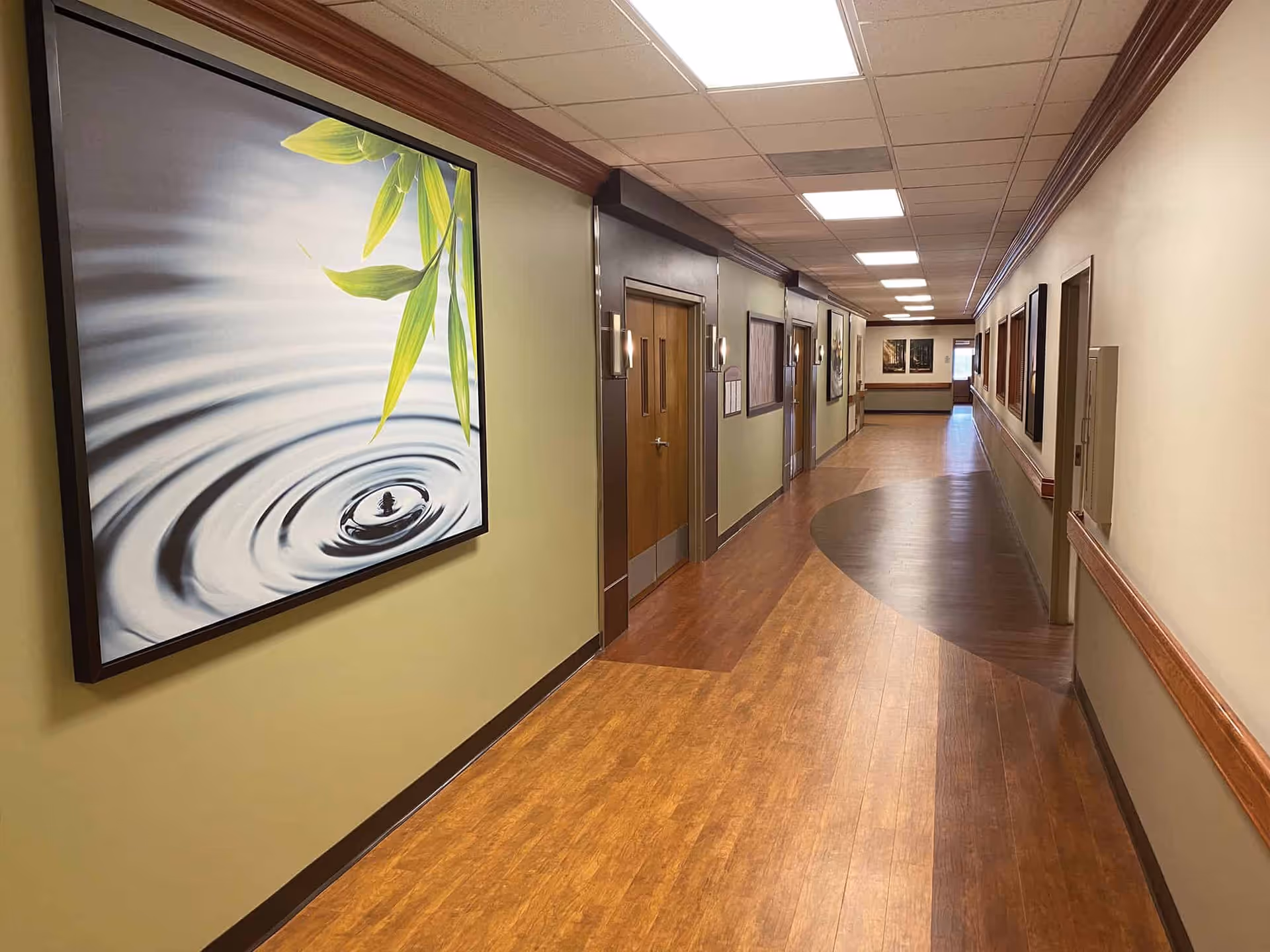 Well-lit, wood-floored interior hallway with framed artwork, doors, and handrails along the walls.