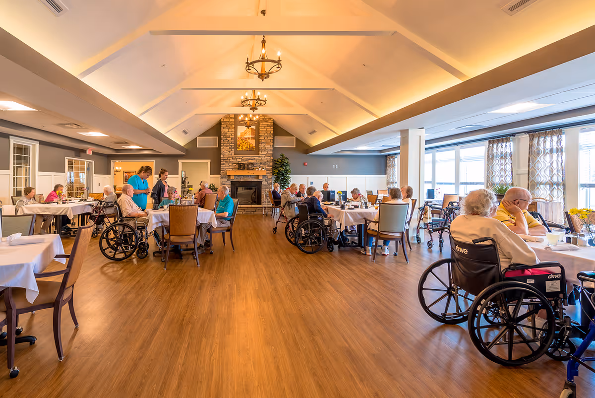 A spacious dining room in a senior living facility with several elderly residents seated at tables, some in wheelchairs, enjoying a meal. The room features a high vaulted ceiling with chandeliers, a stone fireplace at the far end, large windows with patterned curtains, and wooden flooring. Caregivers are assisting some residents.
