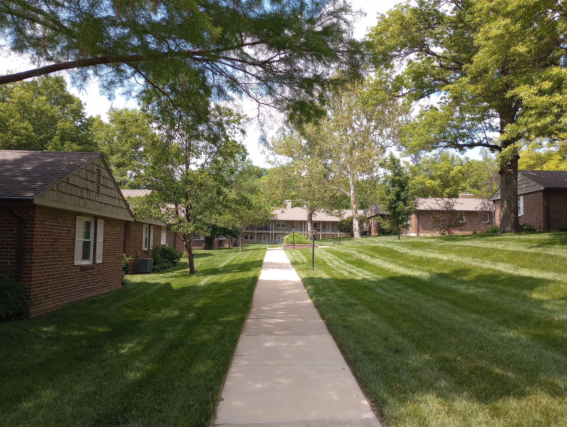 A paved walkway leading through a grassy area with several brick residential buildings on either side, surrounded by trees and greenery under a clear sky.