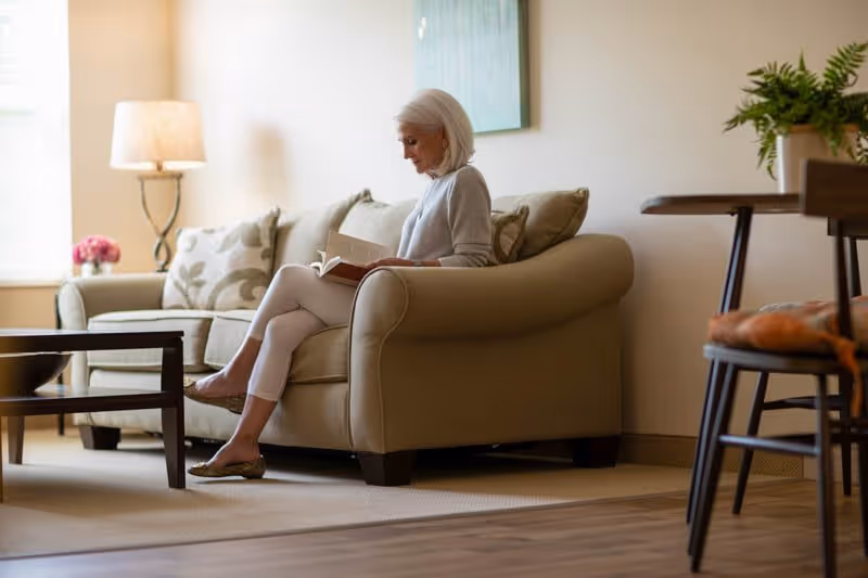An elderly woman with white hair is sitting on a beige sofa in a well-lit living room, reading a book. The room features a floor lamp, a coffee table, a second sofa with patterned cushions, a framed artwork on the wall, and a plant on a table in the background.