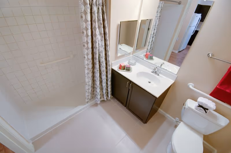 A clean bathroom featuring a white bathtub with a patterned shower curtain, a white sink with a dark wooden cabinet underneath, a large mirror above the sink, and a toilet with a folded towel on top. A red towel hangs on a silver towel rack on the wall.
