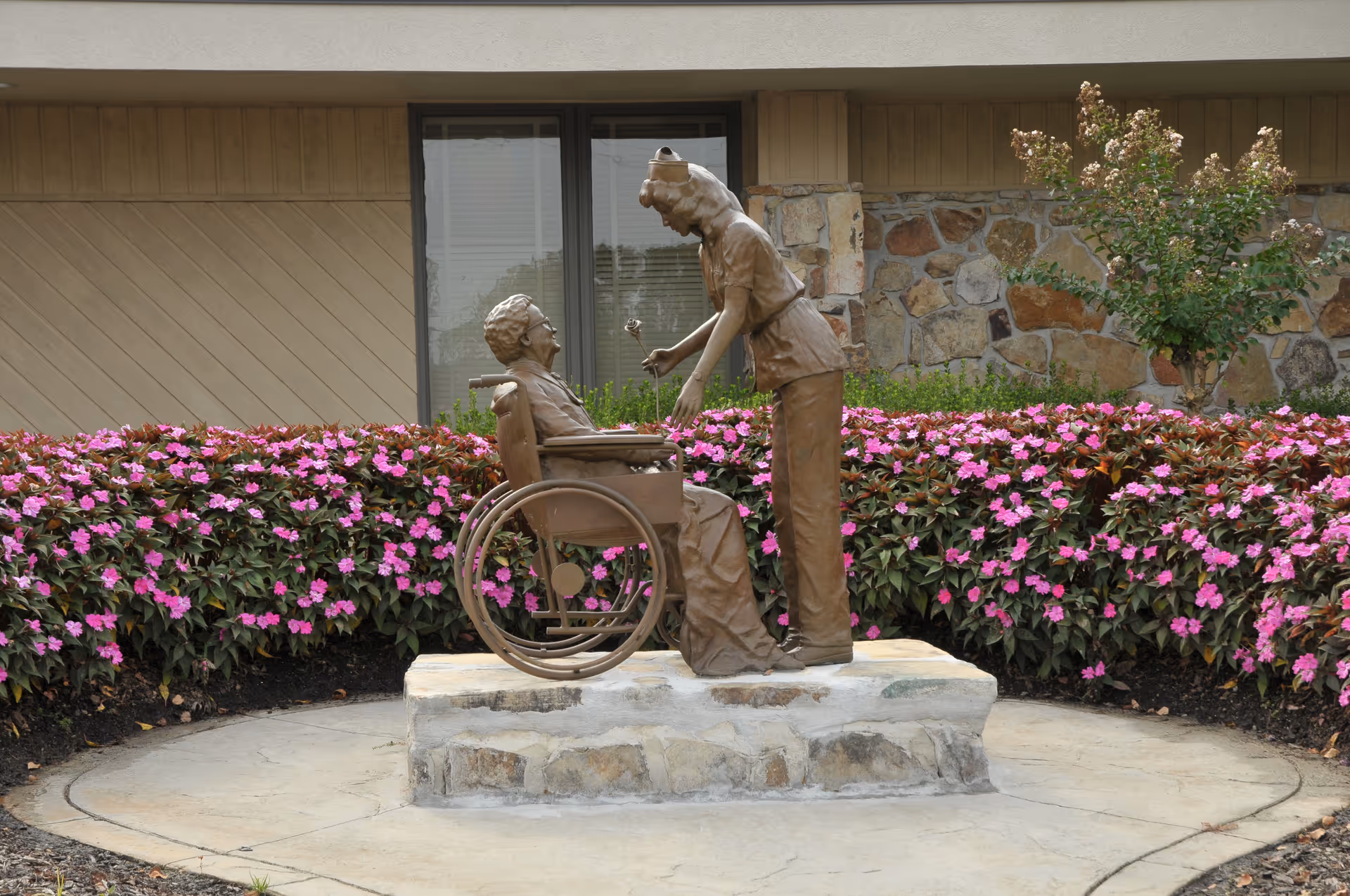 Bronze statue of a caregiver bending over an elderly person in a wheelchair on a stone pedestal surrounded by pink flowers in front of a building.