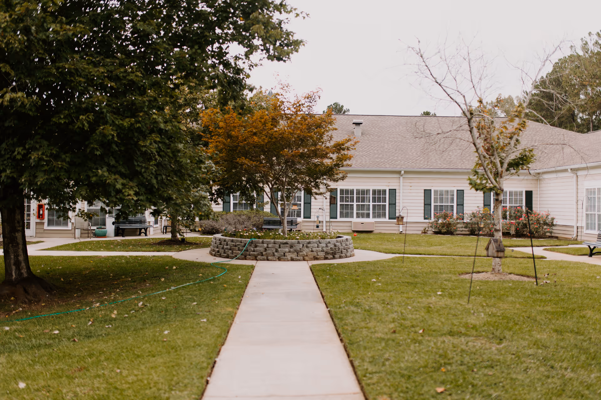 A paved walkway leads through a grassy courtyard with trees and a circular raised flower bed in the center. The courtyard is surrounded by a single-story building with white siding, green shutters, and multiple windows. Benches and bird feeders are visible around the courtyard.