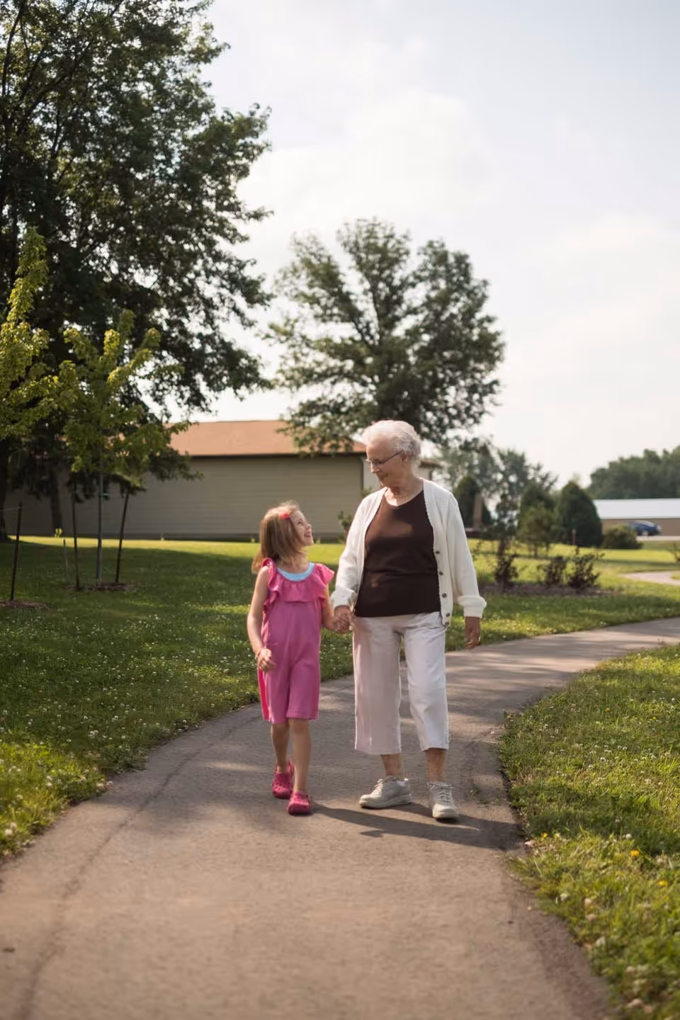An elderly woman and a young girl holding hands and walking together on a paved path in a green outdoor area with trees and buildings in the background.