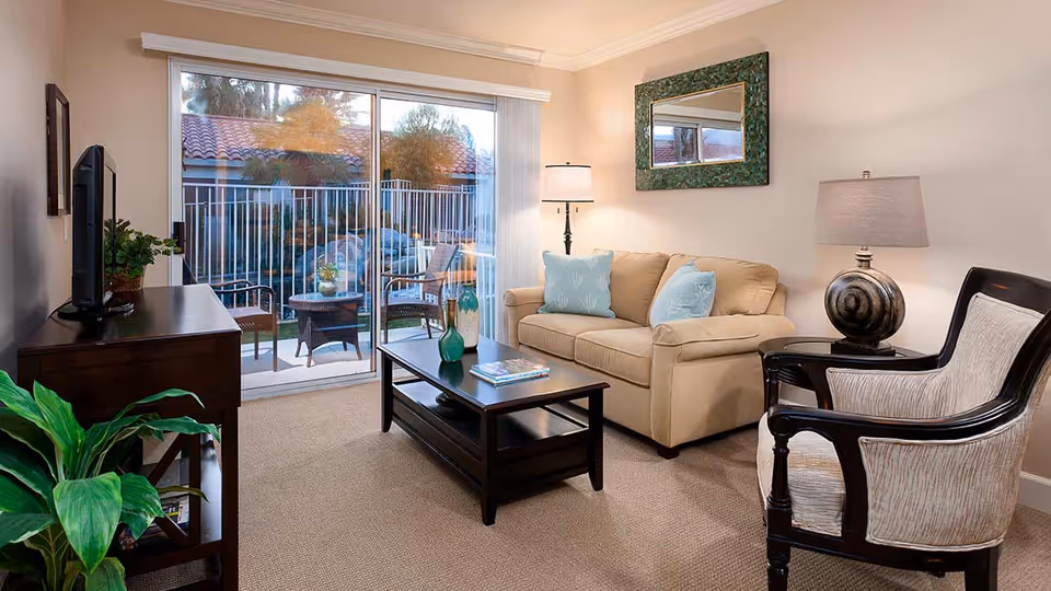 A cozy living room with a beige sofa adorned with light blue pillows, a dark wooden coffee table with a green vase and magazines, a matching dark wooden TV stand with a flat-screen TV, a cushioned armchair with dark wooden frame, two table lamps, a green-framed mirror on the wall, and a sliding glass door leading to an outdoor patio with chairs and a table.