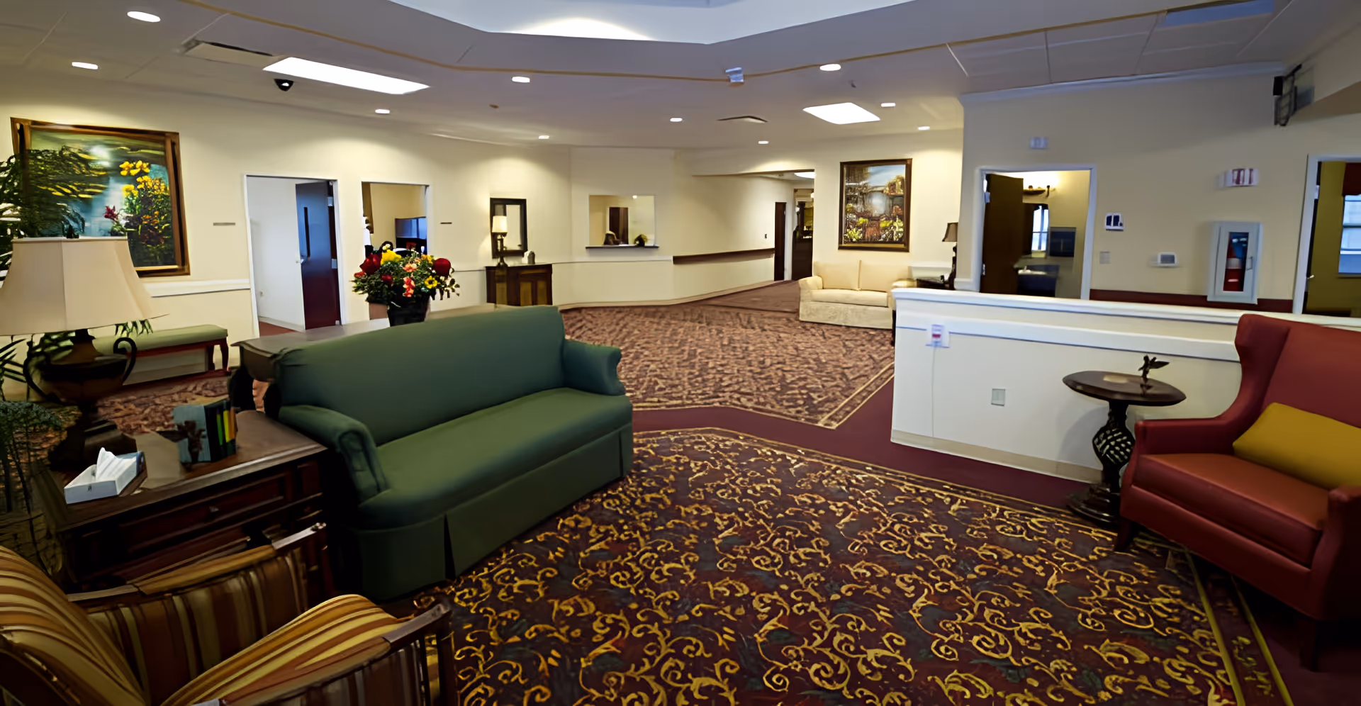 A spacious senior living facility common area with patterned carpet, a green sofa, a red armchair with a yellow cushion, a striped chair, and wooden side tables. The walls are light-colored with framed paintings and several doorways leading to other rooms. The ceiling has recessed lighting and a skylight.