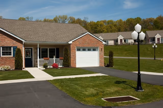 Single-story brick residential building with a front porch, white railing, and a garage. The building is surrounded by a well-maintained lawn and paved driveway. In the background, there are similar buildings and green trees under a clear sky.