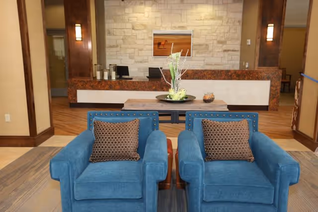 A cozy seating area with two blue armchairs featuring patterned brown cushions, positioned in front of a wooden table with a decorative centerpiece. Behind the seating area is a reception desk with a stone wall backdrop and a framed picture hanging above it. The space has warm lighting and wooden accents.