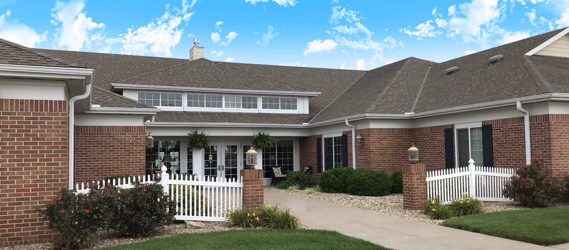 Brick single-story senior living facility entrance with a white picket fence, walkway, shrubs and double glass doors under a blue sky.