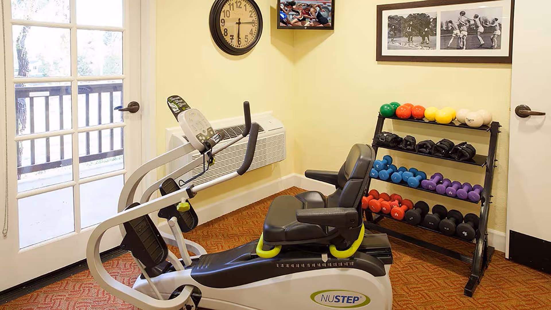 A small exercise room with a NuStep recumbent cross trainer, a rack of colorful dumbbells and weights, a wall clock, framed black and white photos, and a glass door leading outside.