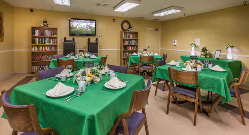 A dining room with several tables covered in green tablecloths, each set with white napkins, plates, cups, and silverware. The room has wooden chairs with purple cushions, bookshelves filled with books, a wall-mounted TV showing a sports game, and decorative flower arrangements on the tables and sideboard against the wall.