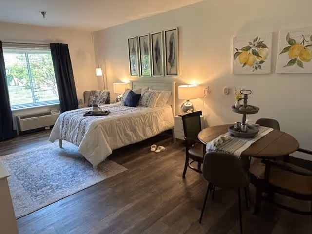 A cozy, well-lit bedroom in an assisted living facility featuring a neatly made bed with white bedding, a small round dining table with four chairs, a floor lamp, and framed artwork on the walls. A window with dark curtains lets in natural light, and a patterned rug is placed under the bed.