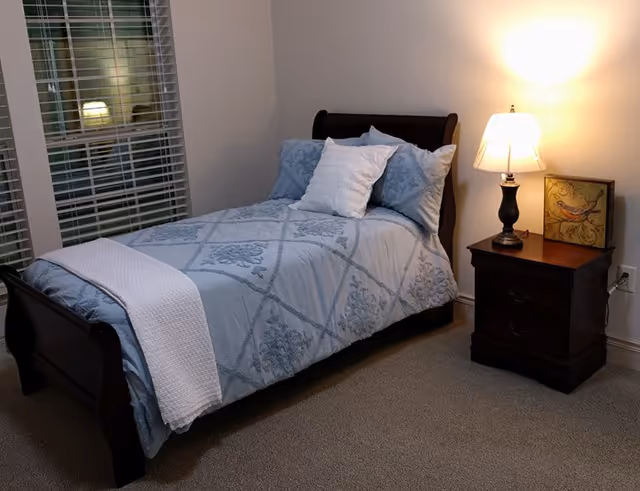 A cozy bedroom with a wooden bed frame featuring a light blue patterned comforter and white pillows. A white throw blanket is draped over the foot of the bed. Next to the bed is a dark wooden nightstand with two drawers, a table lamp with a white shade, and a small framed picture of a bird. The room has beige carpet and white walls, with a window covered by white blinds reflecting the lamp's light.