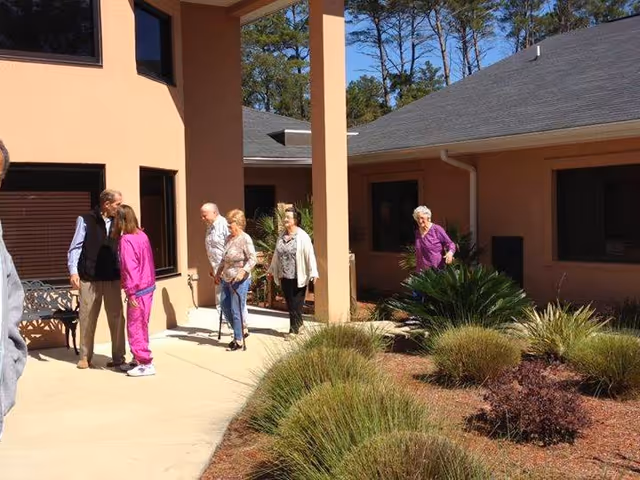 A group of elderly residents walking and talking in a sunny courtyard outside a senior living building.