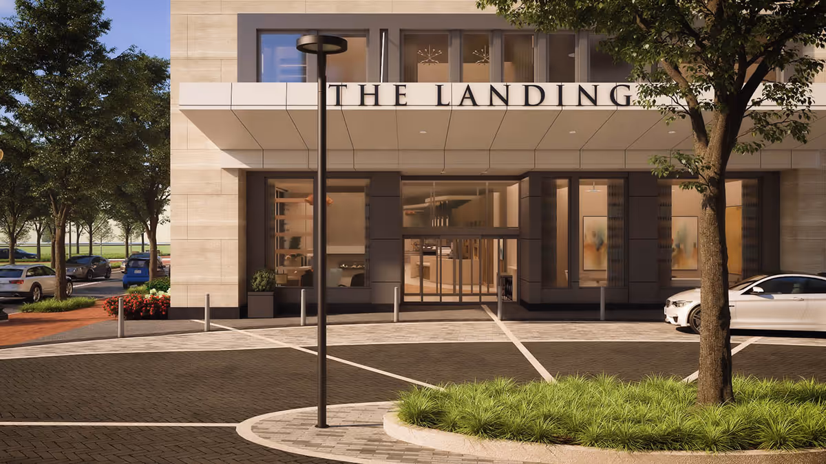 Exterior front entrance of The Landing Alexandria senior living facility with a modern building facade, a tree, landscaped greenery, parked cars, and a streetlamp in the foreground.